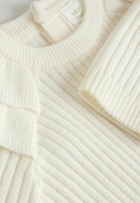 Weißer gerippter Strickbaby-Pullover mit Rüschendetails. Weiche Textur und Rundhalsausschnitt. Etikett innen am Kragen sichtbar.