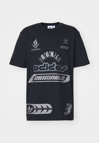 Melna kokvilnas T-kreklā ar lieliem pelēkiem grafiskajiem iespiešanas elementiem, kurā redzams teksts "adidas ORIGINALS", logotipi un stilizēti dizaina elementi. Īsas piedurknes.