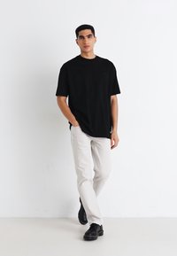 Ung mann står, iført en svart oversized t-skjorte, lysebeige bukser og svarte sko foran en ensfarget hvit bakgrunn.