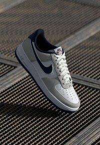Nike Air Force 1 tenisice sa sivo-bijelo-mornarskom bojom, kožnim gornjim dijelom, perforiranim prstom i gumenim đonom.