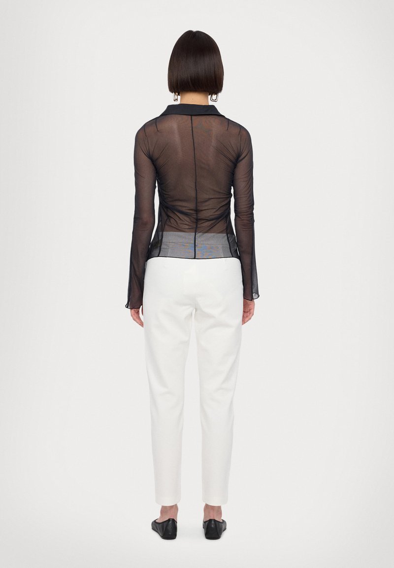 Sort gennemsigtig langærmet top med krave og detaljerede syninger, parret med hvide skræddersyede bukser og sorte flade sko. Minimalistisk design.