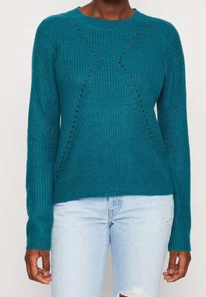 Maglione lavorato a maglia verde acqua con piccoli motivi a rombo traforati, indossato con jeans chiari strappati.