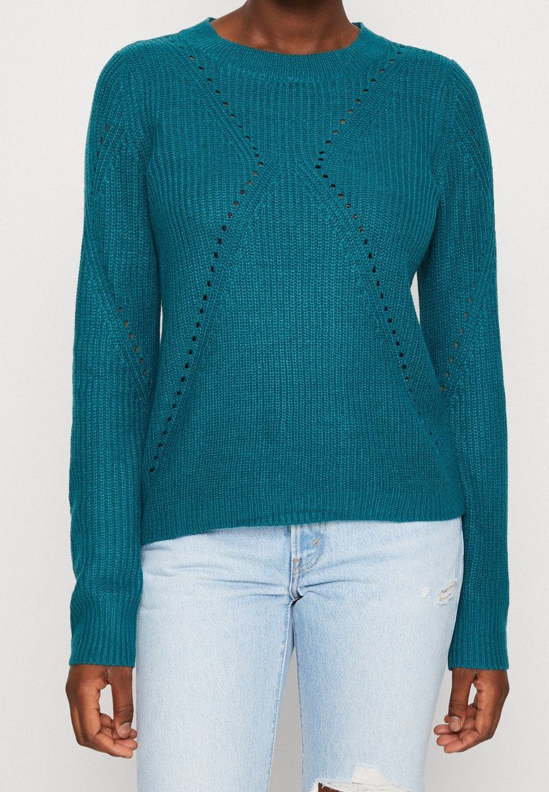 Pull en maille turquoise avec petits motifs losanges ajourés, porté avec un jean bleu clair déchiré.