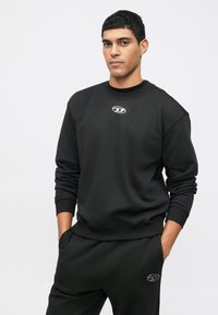 Νεαρός άνδρας με μαύρη φούτερ μπλούζα και παντελόνι που έχει μικρά λευκά λογότυπα, στέκεται με τα χέρια στις τσέπες, μπροστά από φόντο απαλού φωτός.