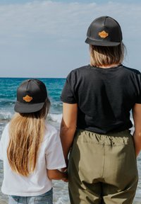 Fekete sapkák narancssárga logóval, felnőtt és gyermek viselésében, fekete pólóval és olajzöld nadrággal párosítva. A háttérben tengerpart és óceán látható.