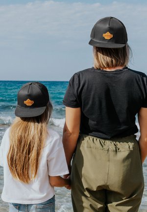Musta lippis, jossa on oranssi logo, päällä aikuisella ja lapsella, yhdistettynä mustaan t-paitaan ja oliivivihreisiin housuihin. Taustalla ranta ja meri.