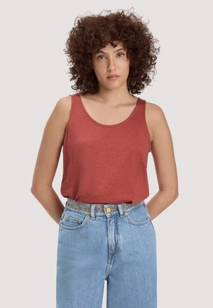 Jonge vrouw met krullend haar, die een mouwloze top in roestkleur draagt, ingestopt in een hoogwater lichtblauwe spijkerbroek met een riem met patroon.