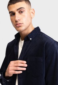 Marineblaue Kordhemd mit Knopfkra=>ger, einzelner Brusttasche und strukturierten vertikalen Linien. Das Model hält den Kragen des Shirts und trägt ein weißes T-Shirt darunter.