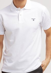 Homme portant un polo blanc uni à manches courtes avec un col et le logo GANT sur la poitrine gauche, main dans la poche.