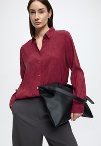Bordeauxrood gestreepte blouse met knopen aan de voorkant en kraag, gecombineerd met grijze broek; houdt een zwarte leren tas met een riem vast.