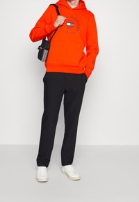 Oranje hoodie met grafische print, zwarte broek en witte sneakers. De hoodie heeft een kangoeroezak en geribbelde manchetten.