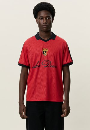 Ung mand med kort krøllet hår iført en rød sportstrøje med sort krave og "Les Deux"-logo, stående mod en ensfarvet lyse baggrund.