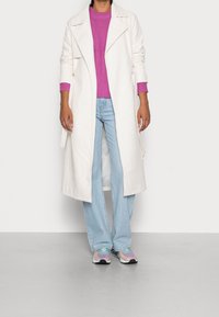 Lång vit kappa över en rosa texturerad tröja, parat med ljusblå utsvängda jeans och flerfärgade sneakers. Slät tyg, moderne design.