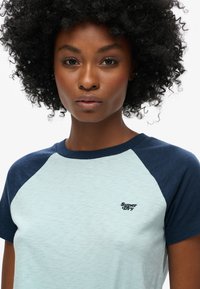 Jasnozielona koszulka z krótkim rękawem, z granatowymi rękawami raglanowymi i teksturowanym wykończeniem, z małym czarnym logo "Superdry" na piersi.