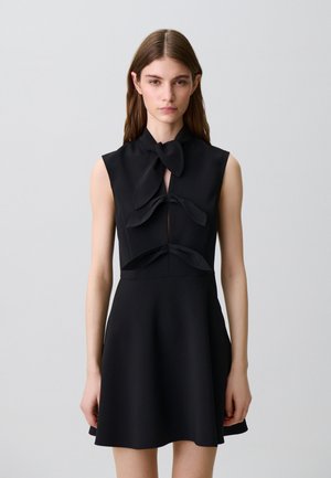 Joven mujer vestida con un vestido negro sin mangas con tres lazos atados en la parte delantera, de pie contra un fondo liso de color gris claro.