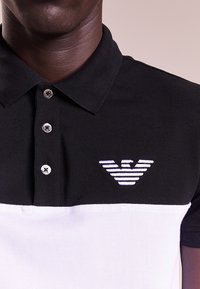 Svart och vit polo med knappad krage, prydd med en randig logotyp broderad på bröstet. Tyget ser texturerat och hållbart ut.