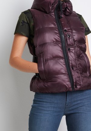 Person iført en mørkelilla dunvest over en kortærmet camouflage-skjorte, hænder i vestens lommer, kombineret med blå jeans.