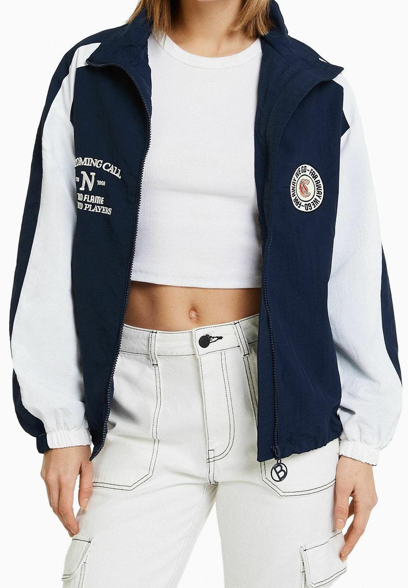 Navy and white windbreaker jacket with elastic cuffs, front zipper, and logo details. Worn over a white cropped shirt and light-colored pants.