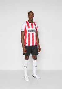 Rød- og hvidstribet fodboldtrøje med sorte detaljer, parret med sorte shorts, hvide sokker og hvide fodboldstøvler med sort logo.