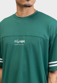 Grønn teksturert mesh t-skjorte med hvite aksertsstriper på ermene. Har «Highlight. REDEFINED REBEL» logo i hvitt på forsiden.