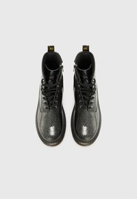 Botas negras con un acabado brillante y brillante, que cuentan con un diseño de tobillo alto, cordones y una suela resistente. Etiqueta de marca amarilla notable en la parte posterior.