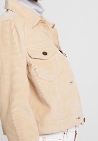 Veste en velours côtelé beige avec un devant boutonné, deux poches poitrine et un design court. Des nervures texturées et des accents en métal sont visibles.