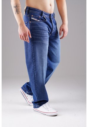 Blaue Jeans aus Denim mit geradem Schnitt, ausgestattet mit Vordertaschen mit sichtbaren Nähten und Kupferbeschlägen. Getragen mit weißen Sneakern.