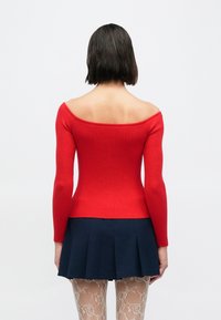 Czerwony prążkowany sweter z odkrytymi ramionami i długimi rękawami, zestawiony z ciemnoniebieską plisowaną spódnicą oraz wzorzystymi kabaretkami.