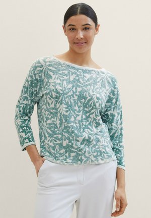 Long-sleeve top featuring a green and white leaf pattern. Made of soft fabric, with a wide neckline and relaxed fit. Paired with white trousers.