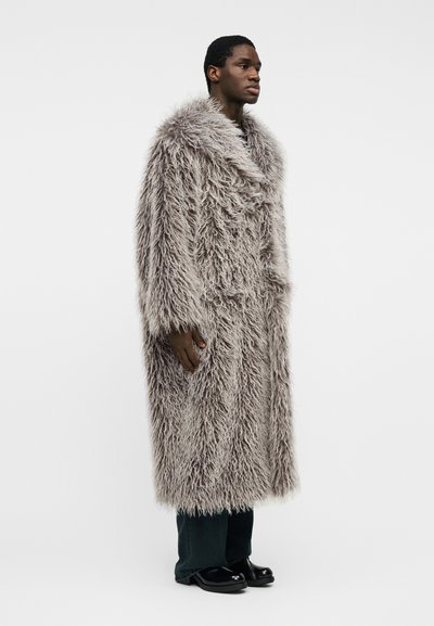 Άνδρας σε πλαϊνή στάση, φοράει ένα μακρύ, γκρίζο, χνουδωτό παλτό από γούνα, σκούρα παντελόνια και μαύρα παπούτσια, μπροστά σε ένα απλό λευκό φόντο.