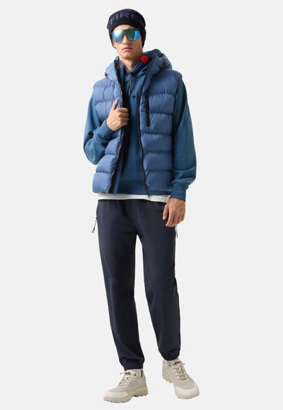 Blaues Steppwesten über einem blauen Hoodie, dunkelblauen Hose und beigen Sneakern. Die Weste hat genähte Abschnitte und eine Kapuze. Die Accessoires bestehen aus einer schwarzen Mütze und einer verspiegelten Sonnenbrille.