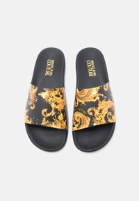 Sandálias tipo slide pretas com um padrão em espiral dourado brilhante. Apresentam uma sola texturada preta e a marca "Versace Jeans Couture" na faixa.