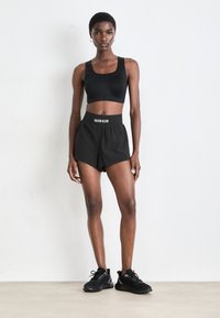 Mujer vestida con un sujetador deportivo negro, pantalones cortos negros de Calvin Klein y zapatillas negras, de pie contra una pared blanca.