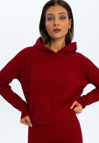 Czerwona bluzka z kapturem z prążkowanej tkaniny, z długimi rękawami, luźnym krojem oraz przednią kieszenią kangurką. Posiada duży kaptur i gładką fakturę.