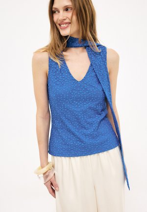 Kvinde iført en ærmeløs blå blomstret tekstureret top med et matchende tørklæde om halsen og cremefarvede bukser.