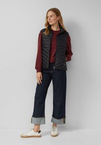 Donna in jeans blu scuri con risvolto, sneakers bianche, maglietta bordeaux a maniche lunghe e gilet smanicato nero trapuntato, in piedi contro un muro bianco.