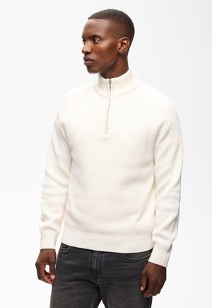 Homme portant un pull en côtes blanc avec une courte fermeture éclair au col et un jean gris foncé, regardant vers sa gauche.