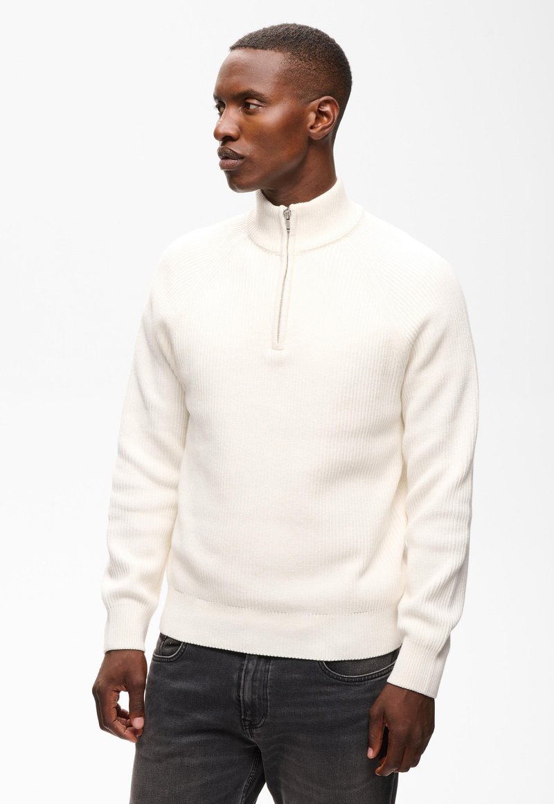 Homme portant un pull en côtes blanc avec une courte fermeture éclair au col et un jean gris foncé, regardant vers sa gauche.