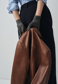 Brown leather jacket held in hands wearing gray knit gloves. Features smooth texture and stitching detail. Striped blue and white shirt visible.