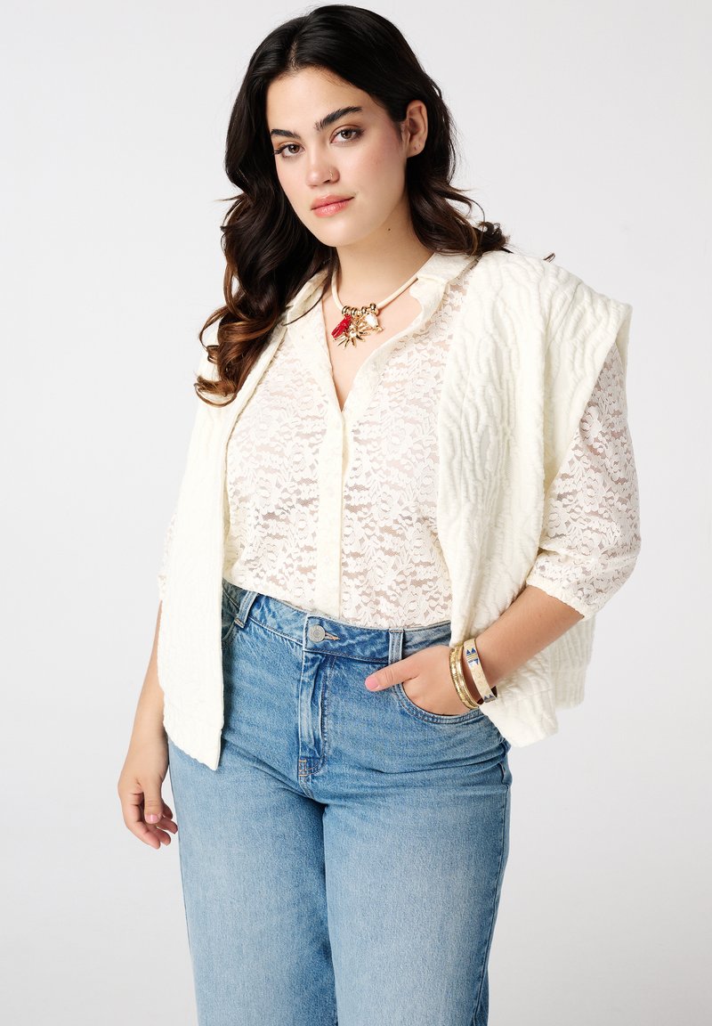 Een witte kantblouse, gelaagd onder een getextureerde crèmewitte vest, gecombineerd met high-waisted blauwe denim, en voorzien van gouden armbanden en een statement ketting.