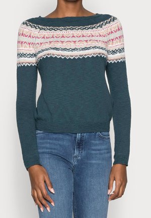 Personne portant un pull en maille bleu sarcelle avec des accents blancs, roses et noirs à motifs sur la poitrine et les épaules, et un jean en denim bleu.