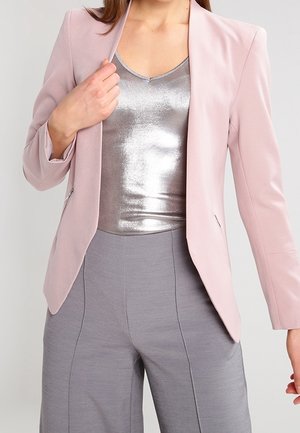 Woman wearing light pink blazer, metallic silver top, and high-waisted gray pants, holding blazer lapel with left hand.