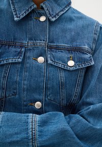 Veste en denim bleu moyen avec un devant à boutons, deux poches poitrine avec rabats et des boutons de couleur argentée. Tissu texturé avec des coutures bien visibles.