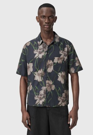 Schlanker Mann trägt ein kurzärmliges, dunkles Hemd mit floralen Mustern, beige Blumen und grünen Stielen, kombiniert mit schwarzen Hosen.