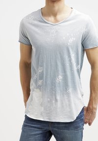 Camiseta de algodón azul claro con escote en pico, que presenta un patrón descolorido y envejecido con manchas blancas, y el dobladillo es más largo en la parte posterior.