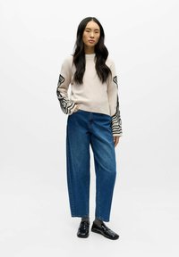 Hellbeiger Pullover mit abstraktem schwarzem Design an den Ärmeln, kombiniert mit hoch taillierten blauen Jeans mit weitem Bein und schwarzen Loafers.