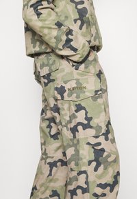 Camouflage-Cargohosen in Grün, Beige und Schwarz. Verfügen über mehrere Taschen und das "BURTON"-Logo in Gold auf der Gesäßtasche.