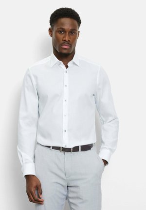 Man draagt een wit overhemd met lange mouwen, ingestopt in lichtgrijze broek met een zwarte riem, staand tegen een witte achtergrond.