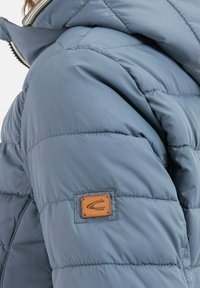 Polstret jakke i lyseblå, med quiltet design, høj krave og et brunt læderlogo på den øverste arm.