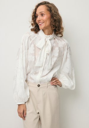 Vrouw met krullend haar die een witte gestructureerde blouse met een strik en beige hoge taille broek draagt, glimlachend en naar de zijkant kijkend.